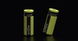 Two yellow cylindrical water bottles with black lids and vertical black stripes featuring the word 'AQUA' are placed against a dark background. One bottle stands upright while the other is slightly tilted.