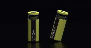Two yellow cylindrical water bottles with black lids and vertical black stripes featuring the word 'AQUA' are placed against a dark background. One bottle stands upright while the other is slightly tilted.