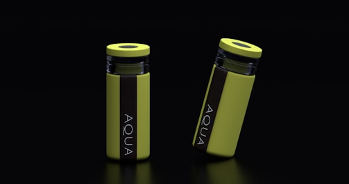 Two yellow cylindrical water bottles with black lids and vertical black stripes featuring the word 'AQUA' are placed against a dark background. One bottle stands upright while the other is slightly tilted.