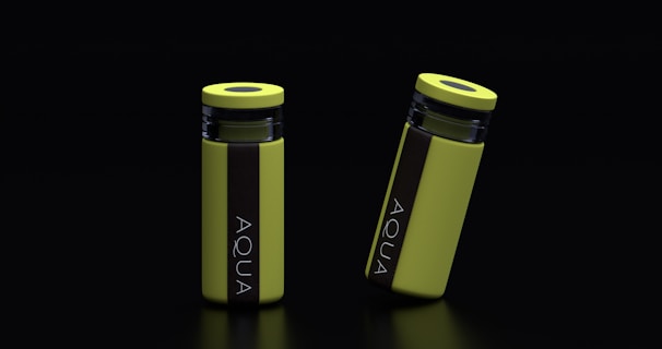Two yellow cylindrical water bottles with black lids and vertical black stripes featuring the word 'AQUA' are placed against a dark background. One bottle stands upright while the other is slightly tilted.