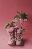 A pair of beige sneakers is filled with vibrant pink flowers and green leaves, set against a matching pink background. The flowers spill over the tops of the shoes, creating an artistic and whimsical arrangement.