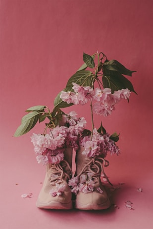 A pair of beige sneakers is filled with vibrant pink flowers and green leaves, set against a matching pink background. The flowers spill over the tops of the shoes, creating an artistic and whimsical arrangement.
