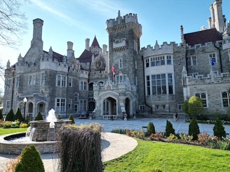 A large, historic castle featuring a stone facade, multiple turrets, and intricate architectural details. A fountain is situated in the foreground, surrounded by manicured gardens with neatly trimmed hedges and pathways. The sky is clear and blue, casting bright sunlight on the scene.