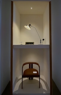 A minimalist display cabinet containing a modern chair, a vintage typewriter, and a desk lamp. The lamp is positioned above, casting soft light over the typewriter and the chair below. The setting is simple, with a clean white backdrop and wooden borders framing the space. There is an ambient light from above, creating a focused and intimate atmosphere.