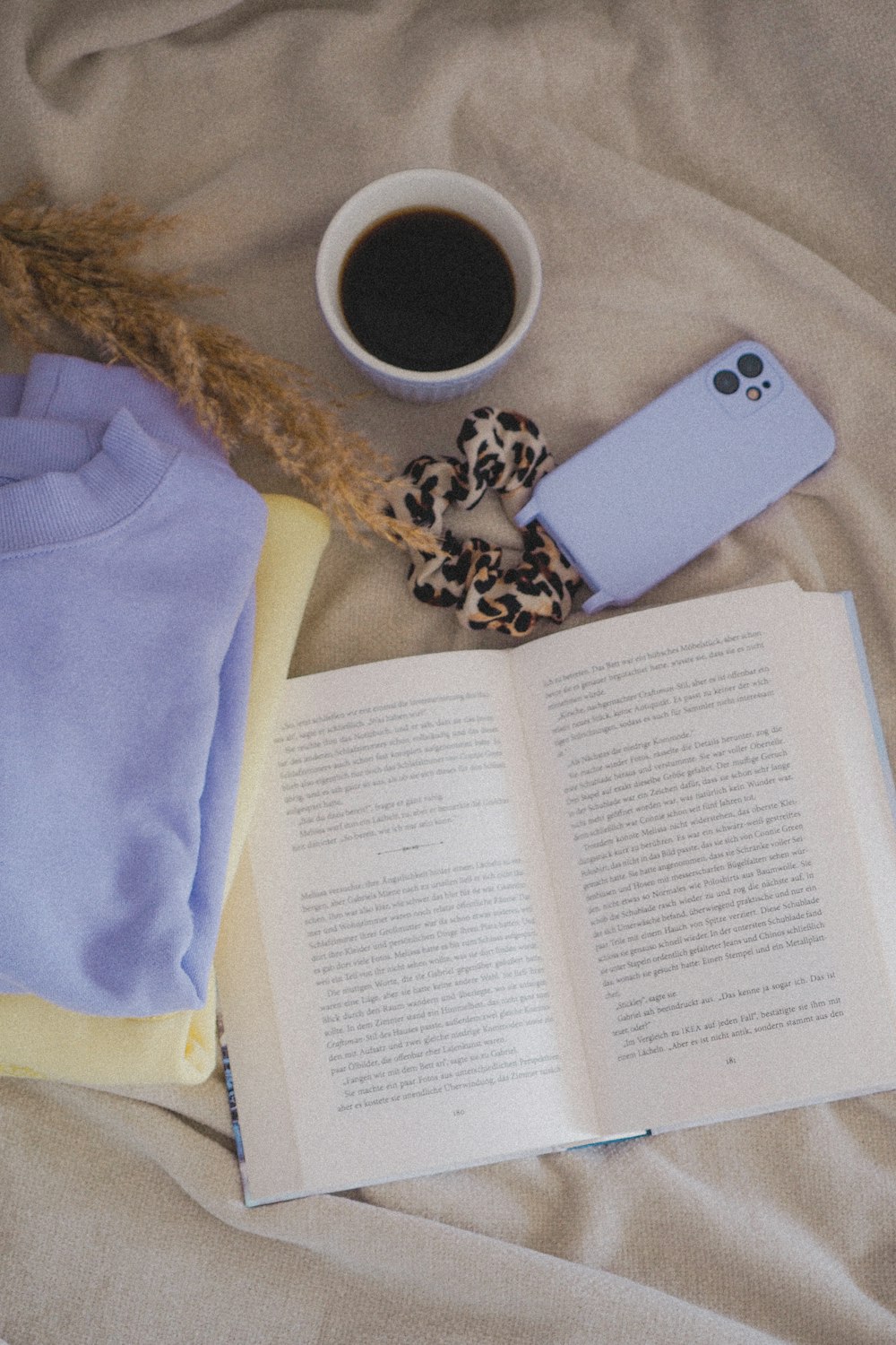 a cup of coffee, a cell phone, and an open book on a bed