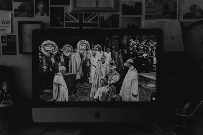 A black and white image of a Mac monitor displaying a historical or ceremonial event with several people dressed in ornate robes, possibly from a coronation or similar occasion. In the background, there are various framed pictures on the wall, including photographs of people and a Union Jack flag.