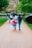 People walking down a park path on a rainy day. One individual is wearing a rain poncho with a Union Jack flag design. Several people are holding black umbrellas, and the path is lined with green grass and trees.
