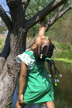 A child in a green dress is playfully hanging onto a tree branch. The child has braided hair adorned with green and white beads. The background features a lush, natural setting with a body of water and greenery.