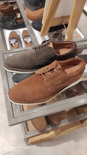 A pair of casual lace-up shoes displayed on a glass shelf in a shoe store. One shoe is brown and the other is dark brown, showcasing a textured material. In the background, additional pairs of shoes are visible, including slip-ons and loafers, all arranged neatly on shelves.
