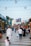A busy outdoor market scene with various stalls along a wide walkway. People of different ages and backgrounds are walking and browsing. Overhead, strings of lights and star decorations enhance the lively ambiance. The market is bustling with activity as shoppers explore stalls displaying colorful goods.