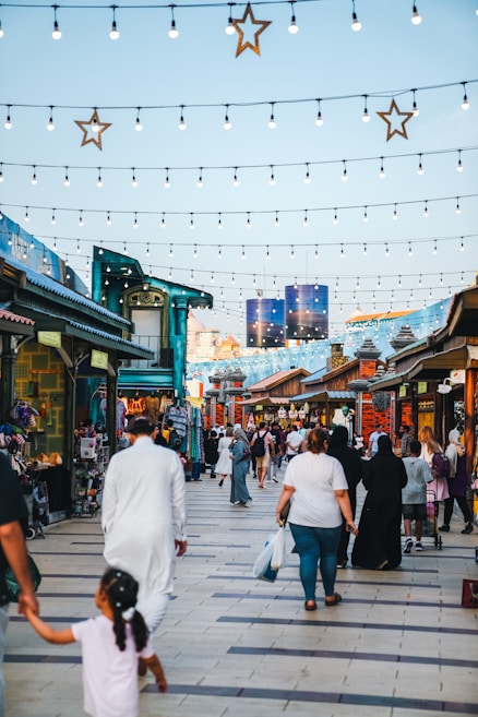 A busy outdoor market scene with various stalls along a wide walkway. People of different ages and backgrounds are walking and browsing. Overhead, strings of lights and star decorations enhance the lively ambiance. The market is bustling with activity as shoppers explore stalls displaying colorful goods.
