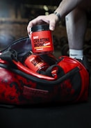 A hand reaches into a red and black gym bag to pick up a container labeled 'Sweoly Creatine Monohydrate'. The gym bag and container are prominently red with black detailing. The container appears to be for a fitness supplement, and there is another similar container partially visible inside the bag.