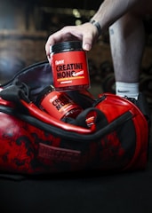 A hand reaches into a red and black gym bag to pick up a container labeled 'Sweoly Creatine Monohydrate'. The gym bag and container are prominently red with black detailing. The container appears to be for a fitness supplement, and there is another similar container partially visible inside the bag.
