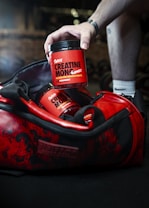 A hand reaches into a red and black gym bag to pick up a container labeled 'Sweoly Creatine Monohydrate'. The gym bag and container are prominently red with black detailing. The container appears to be for a fitness supplement, and there is another similar container partially visible inside the bag.
