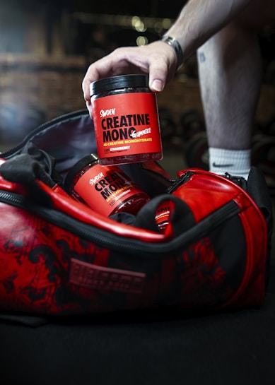 A hand reaches into a red and black gym bag to pick up a container labeled 'Sweoly Creatine Monohydrate'. The gym bag and container are prominently red with black detailing. The container appears to be for a fitness supplement, and there is another similar container partially visible inside the bag.