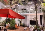 An outdoor patio area featuring a large red umbrella, surrounded by trees and plants. Several chairs and a table are positioned on a brick-paved space. String lights hang between trees, and a building with a balcony is in the background. A sign reading 'Wunder Garden' is prominently displayed.