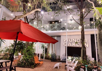 An outdoor patio area featuring a large red umbrella, surrounded by trees and plants. Several chairs and a table are positioned on a brick-paved space. String lights hang between trees, and a building with a balcony is in the background. A sign reading 'Wunder Garden' is prominently displayed.