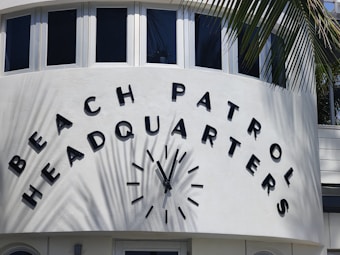 A curved building facade with large letters spelling out 'Beach Patrol Headquarters.' A clock is centered between the words, and palm fronds cast shadows on the white wall.