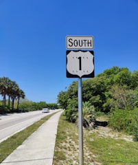 A well-maintained road sign indicating a southward direction on Route 1, with a clear blue sky in the background. The scene includes a road bordered by lush green trees and bushes on the right, and palm trees on the left. A couple of cars are visible driving along the road.