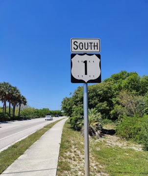 A well-maintained road sign indicating a southward direction on Route 1, with a clear blue sky in the background. The scene includes a road bordered by lush green trees and bushes on the right, and palm trees on the left. A couple of cars are visible driving along the road.