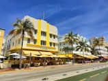 Art Deco buildings with pastel colors and yellow accents line a sunny street, adorned with palm trees and outdoor cafes. People are seated under umbrellas enjoying the day. The sky is a clear blue, adding to the lively and relaxed atmosphere.