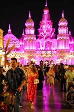 A vibrant, illuminated building showcases intricate architectural details with pink and purple lighting at night. A diverse crowd of people, including families and individuals, walks along a busy promenade, some taking photos and others casually strolling.