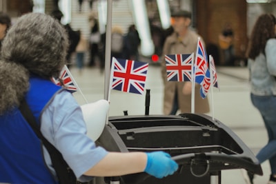 A person wearing a blue uniform and gloves is pushing a cleaning cart adorned with multiple small Union Jack flags. The environment appears to be a busy place, possibly a station or airport, with people in the background walking or standing.