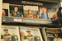A display rack filled with various newspapers and magazines featuring images and articles about King Charles III's coronation. Prominent colors include blues and reds, and the display has a header celebrating the coronation.