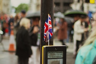 A small British flag is attached to a post near a pedestrian crossing button. The background features a crowd of people standing, many holding umbrellas, suggesting a rainy day. The scene appears to be in an urban setting, with blurred traffic cones visible.
