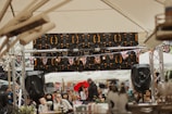 A crowded outdoor event with people gathered under large tents. British flags decorate the area, indicating a possible cultural or national celebration. Audio equipment and speakers are visible, suggesting a performance or speech is ongoing. The attendees appear engaged, and some are holding drinks.