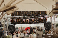A crowded outdoor event with people gathered under large tents. British flags decorate the area, indicating a possible cultural or national celebration. Audio equipment and speakers are visible, suggesting a performance or speech is ongoing. The attendees appear engaged, and some are holding drinks.