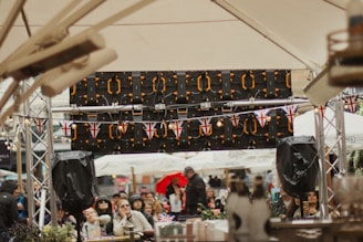 A crowded outdoor event with people gathered under large tents. British flags decorate the area, indicating a possible cultural or national celebration. Audio equipment and speakers are visible, suggesting a performance or speech is ongoing. The attendees appear engaged, and some are holding drinks.