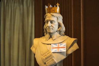 A statue bust of a historical figure wearing a paper crown with the words 'Save the King', and a sign with the British flag reading 'Long Live King Charles' attached to its front. The background features a wooden panel and a curtain.