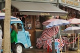 A vibrant street scene features a vintage blue food truck parked against a building with signs reading 'Spital's Wine Bar' and 'The Golden Heart.' Union Jack-themed decorations, including tablecloths, bunting, and umbrellas, are prominently displayed. Two individuals are visible, one woman in a green jacket near the food truck. The setting has a festive atmosphere with outdoor seating.