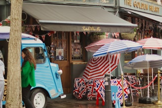 A vibrant street scene features a vintage blue food truck parked against a building with signs reading 'Spital's Wine Bar' and 'The Golden Heart.' Union Jack-themed decorations, including tablecloths, bunting, and umbrellas, are prominently displayed. Two individuals are visible, one woman in a green jacket near the food truck. The setting has a festive atmosphere with outdoor seating.