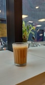A glass of chai sits on a light wooden table near a window. Outside, rain droplets are visible on the glass, and a green plant is in the background. The setting has an indoor-outdoor ambiance with hints of natural and artificial lighting.