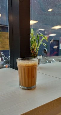 A glass of chai sits on a light wooden table near a window. Outside, rain droplets are visible on the glass, and a green plant is in the background. The setting has an indoor-outdoor ambiance with hints of natural and artificial lighting.