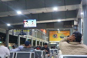 A waiting area in a public transport terminal with several people seated. Overhead, there are signs indicating directions and a television screen displaying a news broadcast. The space is brightly lit with modern design elements, including metal columns and comfortable seating.