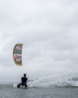 A person engages in kitesurfing on a body of water under a cloudy sky. The colorful kite contrasts with the overcast atmosphere as the surfer skims across the water, creating splashes.