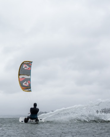 A person engages in kitesurfing on a body of water under a cloudy sky. The colorful kite contrasts with the overcast atmosphere as the surfer skims across the water, creating splashes.
