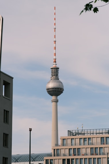 A striking television tower rises against a backdrop of a partly cloudy sky. The tower has a distinctive spherical structure near its top, followed by an antenna extending upwards. Surrounding the tower are modern buildings with flat roofs and large windows.