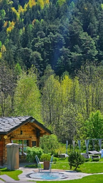 A cozy log cabin nestled in a lush forest setting with a backdrop of dense, green trees. In front of the cabin, there's a circular hot tub surrounded by well-maintained grass and a few loungers, suggesting a serene getaway spot.
