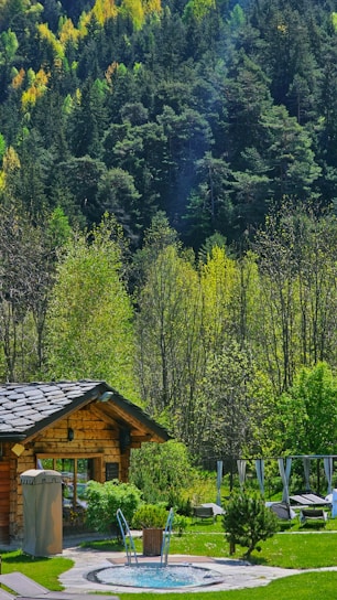 A cozy log cabin nestled in a lush forest setting with a backdrop of dense, green trees. In front of the cabin, there's a circular hot tub surrounded by well-maintained grass and a few loungers, suggesting a serene getaway spot.