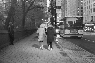 A city street scene at night features two people in winter coats walking hand in hand on a cobblestone sidewalk. They pass by a large parked bus, with buildings and city lights in the background. Another person stands alone by a tree, dressed in a hooded jacket.