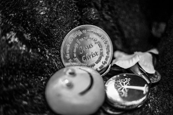 A silver coin featuring the engraved text 'Jesus Christ' lies amidst other small objects, including a round item with a smiley face and a button with a tree design. The background is a textured dark surface, possibly fabric.