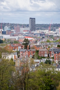 A cityscape image depicting a mix of residential and commercial buildings. In the foreground, there are traditional red-brick houses surrounded by green trees. In the background, modern high-rise buildings and construction cranes are visible. The sky is covered with clouds, suggesting an overcast day.