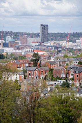 A cityscape image depicting a mix of residential and commercial buildings. In the foreground, there are traditional red-brick houses surrounded by green trees. In the background, modern high-rise buildings and construction cranes are visible. The sky is covered with clouds, suggesting an overcast day.
