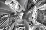 An intricate architectural interior featuring undulating curves, glass panels, and a play of light and shadows. The perspective is angled upward, capturing the dynamic and flowing design elements that create a sense of movement and modernism.