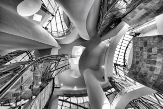An intricate architectural interior featuring undulating curves, glass panels, and a play of light and shadows. The perspective is angled upward, capturing the dynamic and flowing design elements that create a sense of movement and modernism.