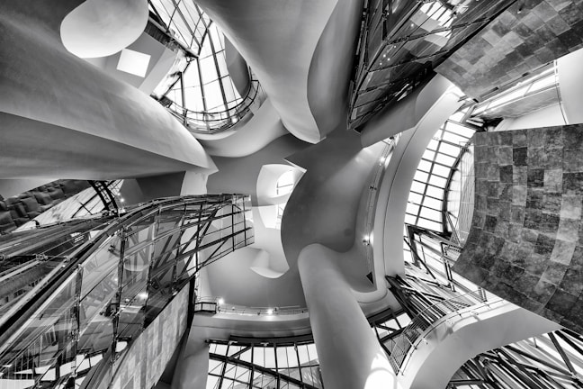 An intricate architectural interior featuring undulating curves, glass panels, and a play of light and shadows. The perspective is angled upward, capturing the dynamic and flowing design elements that create a sense of movement and modernism.